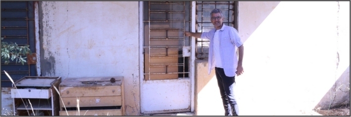 Man in white shirt standing in front of the door to an abandoned white building.