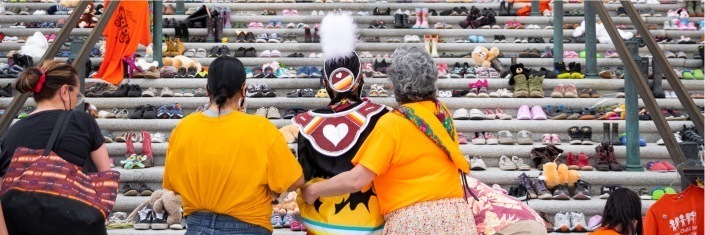 Three First Nations women facing steep set of stairs pairs of children's shoes cover each step