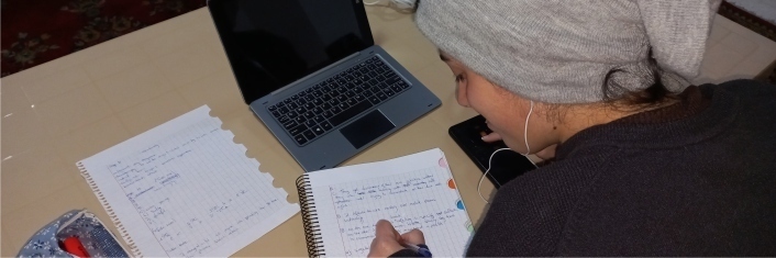 A young woman sitting at a table writing in a booklet with computercalculator